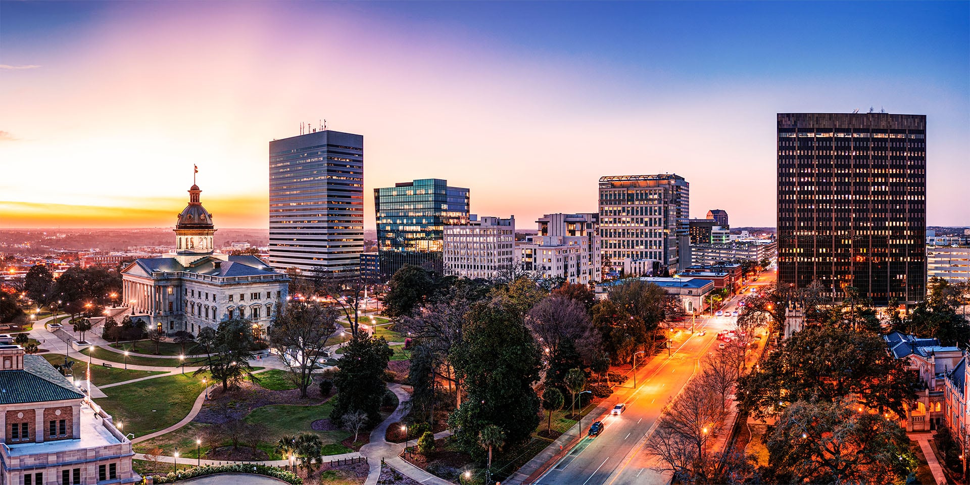 Nighttime view of downtown Columbia, South Carolina, with city lights illuminating the skyline. Tall buildings and the iconic South Carolina State House dome are visible, reflecting a vibrant urban scene under a dark sky